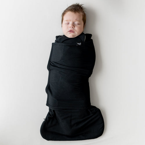 Sleeping infant laying on a light neutral blanket swaddled in the Sleep Bag Swaddler in Midnight 1.0
