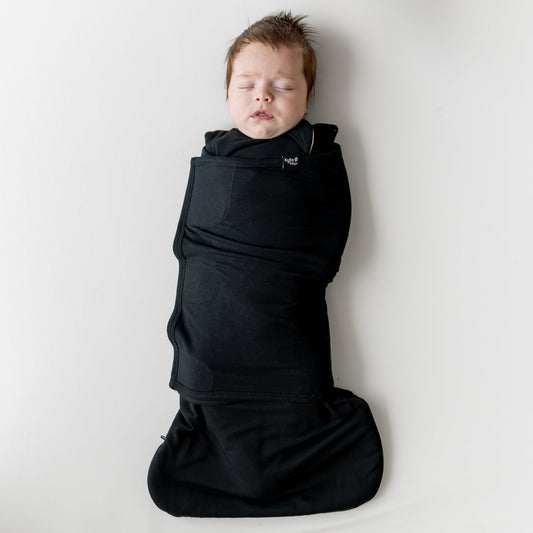 Sleeping infant laying on a light neutral blanket swaddled in the Sleep Bag Swaddler in Midnight 1.0
