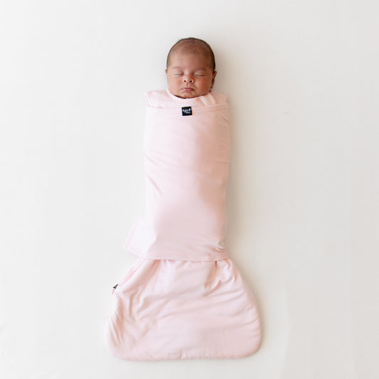 Baby sleeping on a white surface wearing a sleep bag swaddler in a light pink