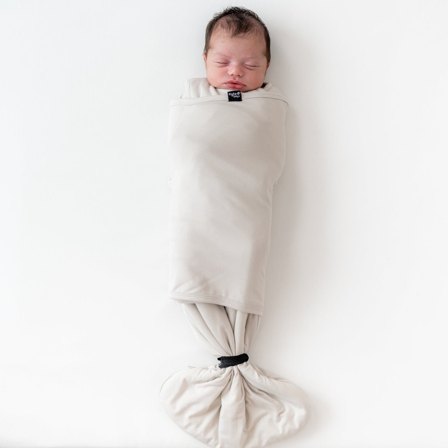 Sleeping baby wearing Kyte Baby Sleep Bag Swaddler in Oat with elastic band at bottom
